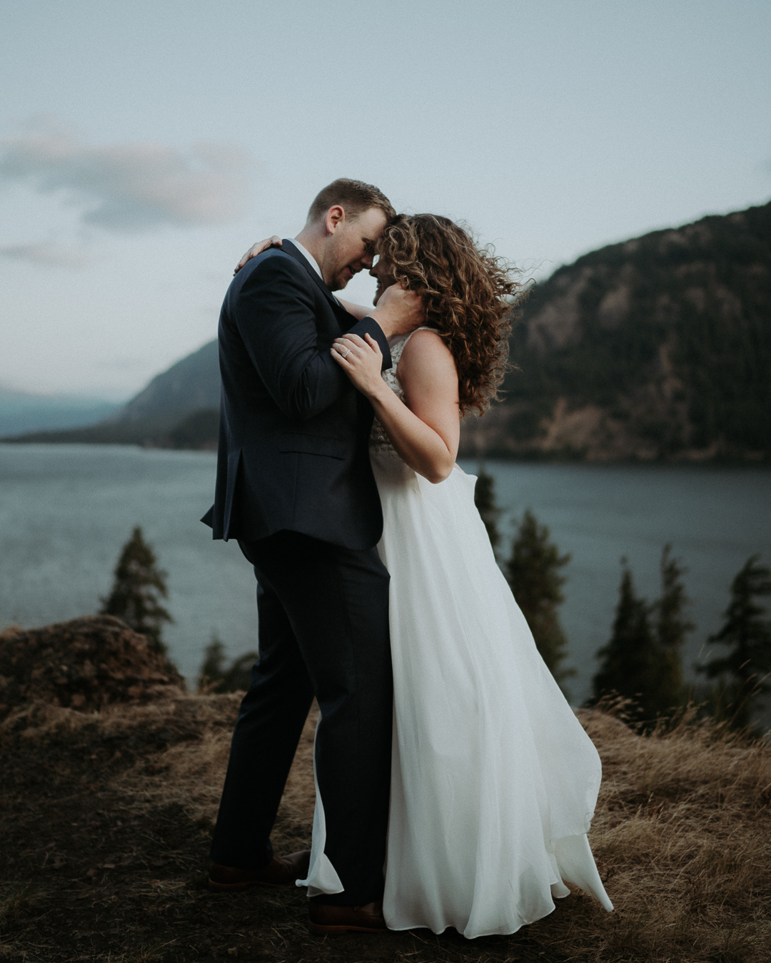 Bryce and Britnay leaning their foreheads together above a river and wooded cliffs
