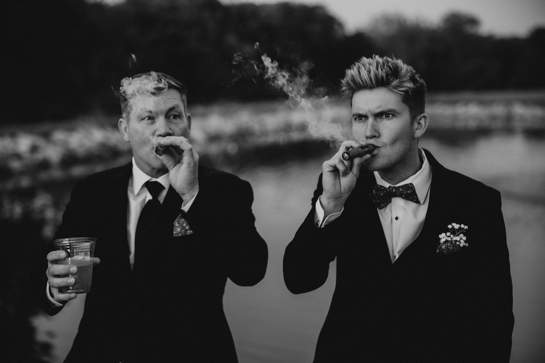 Two men in wedding suits sharing a cigar break in a black-and-white photograph